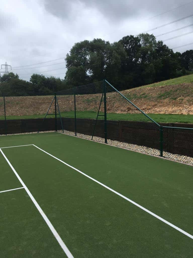 This is a photo of a newly installed tennis court, with a new green metal fence. Behind the metal fence there is a shingle walkway , and there are sleepers that are acting as a retaining wall, as the new court has been cut in to the ground.