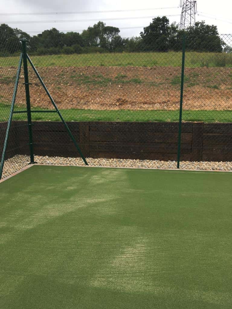 This is a photo of a newly installed tennis court, with a new green metal fence. Behind the metal fence there is a shingle walkway , and there are sleepers that are acting as a retaining wall, as the new court has been cut in to the ground.