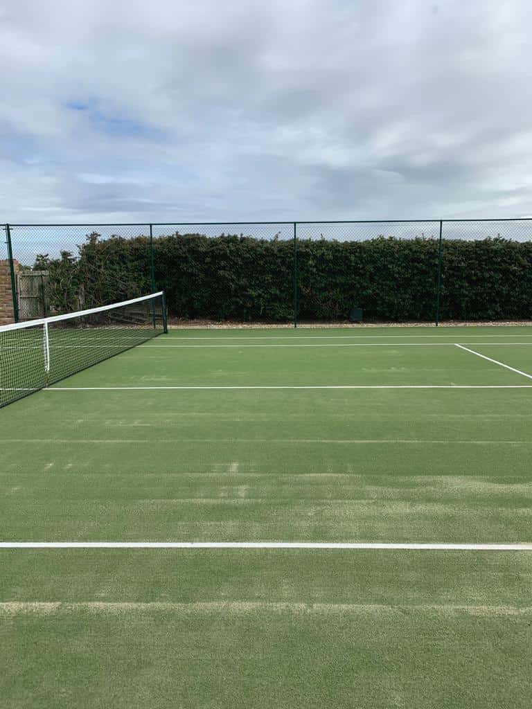 This is a photo of a resurfaced tennis court with a high fence installed, and behind the fence is a large hedge.