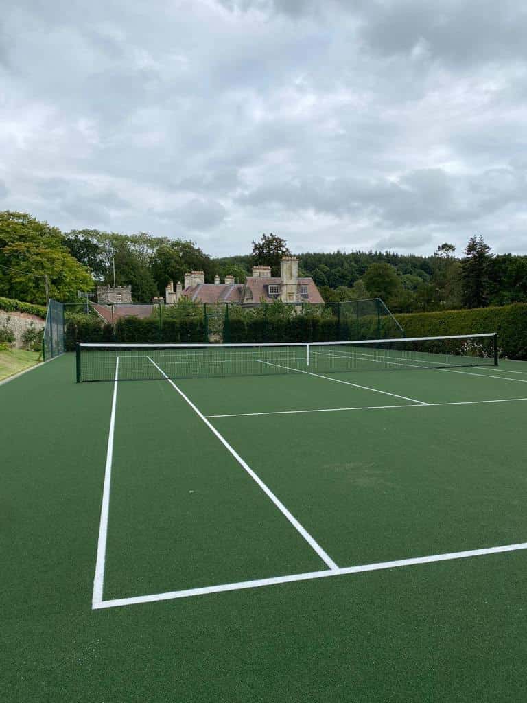 This is a photo of a newly installed tennis court, the court has a drop fence on both sides, one side is enclosed by a hedge. In the distance there is a very large county house and trees.